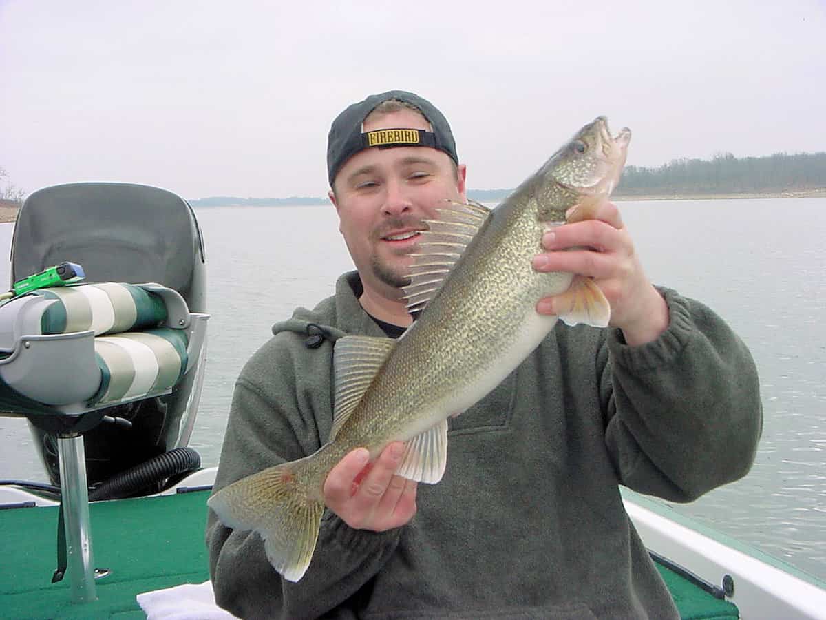 An angler on a boat holds up a nice-sized walleye he caught fishing at Stockton Lake with the water behind him.