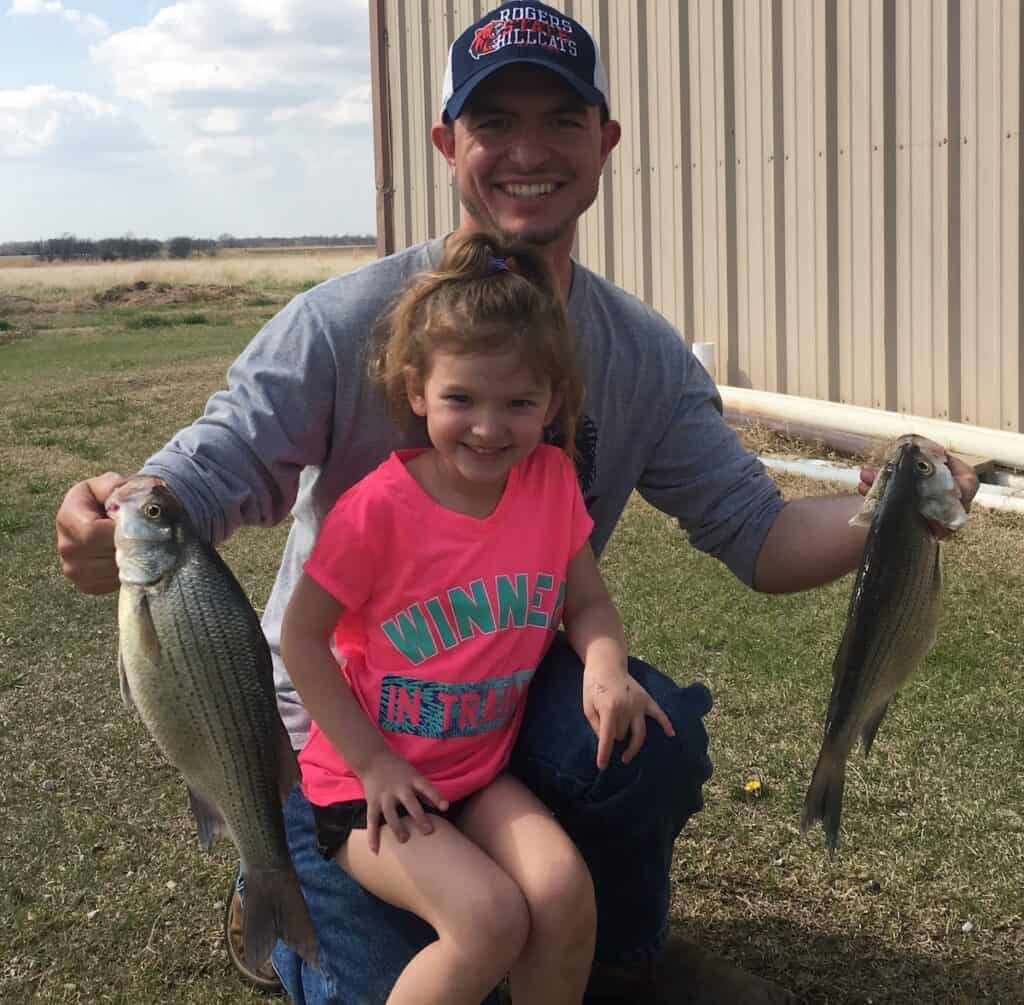 Author Wesley Littlefield kneels holding two white bass caught fishing in Oklahoma, with his daughter kneeling in front of him and a metal sized building in the background.