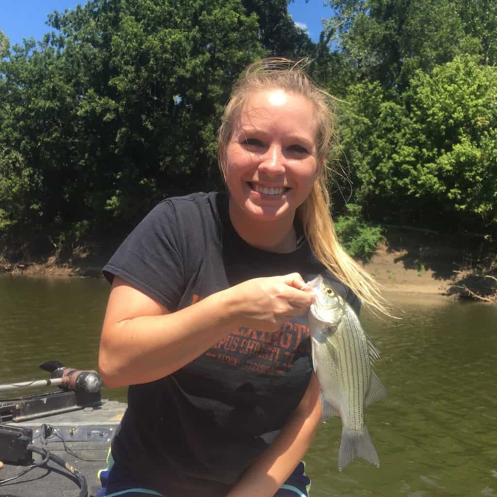 Woman on a boat holds a white bass caught while fishing in Oklahoma, with water and trees in the background.