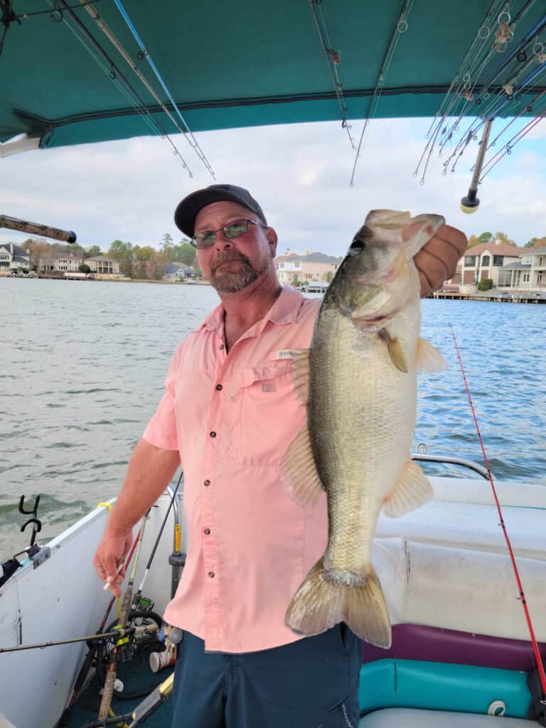 Angler on a boat holds a good-sized largemouth bass by the lip with Lake Conroe and lakeshore houses in the background.