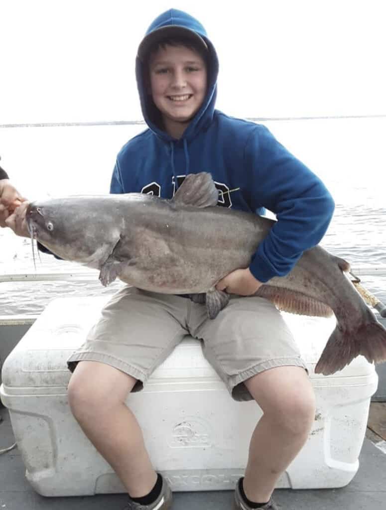 Boy sitting on cooler holds a massive catfish on his lap while riding a boat, with the water of Lake Conroe, Texas, in the background.