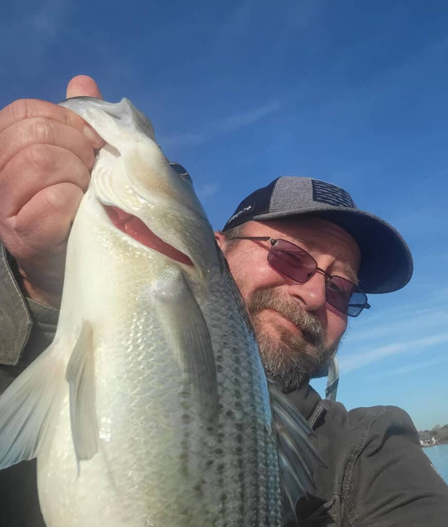 Closeup selfie of a smiling man holding a hybrid striped bass caught fishing in Lake Conroe.