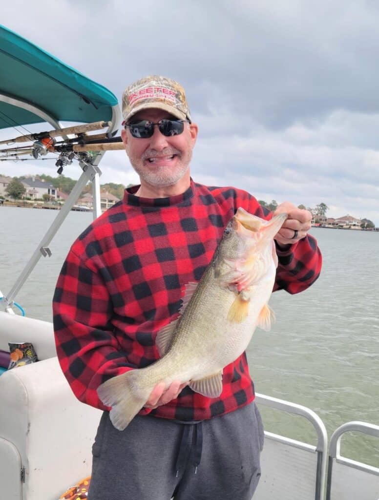 A smiling man in a fishing boat holds a bass he caught at Lake Conroe, which shows in the background.
