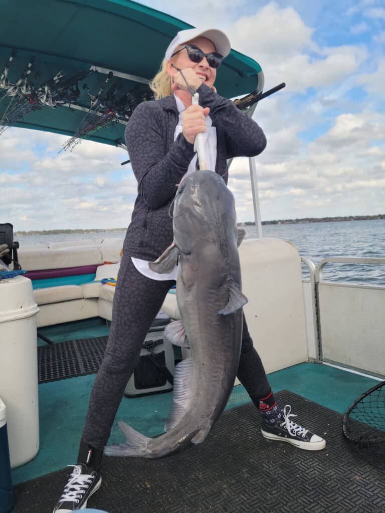 Woman in a boat holds up a giant catfish caught fishing with Lake Conroe in the background.