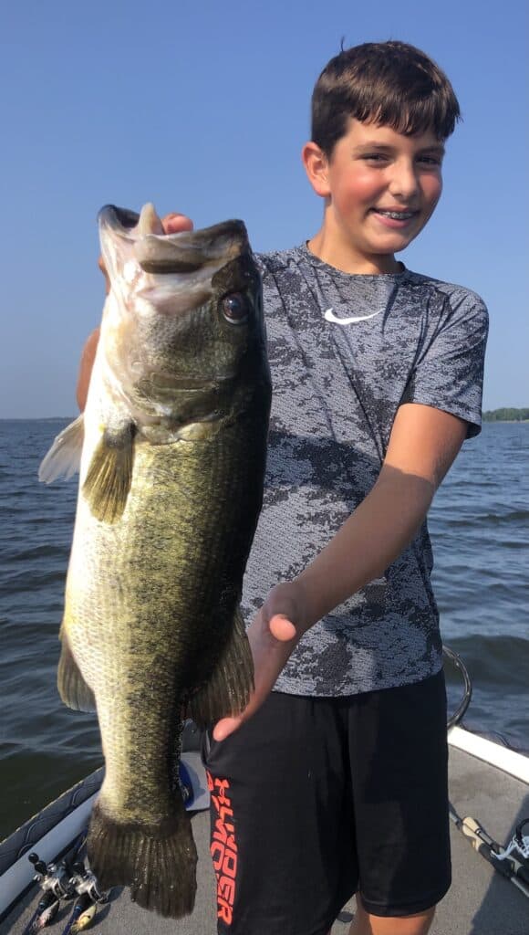 A young, smiling boy in a boat holds up a giant largemouth bass with the water of Lake Fork in the background.