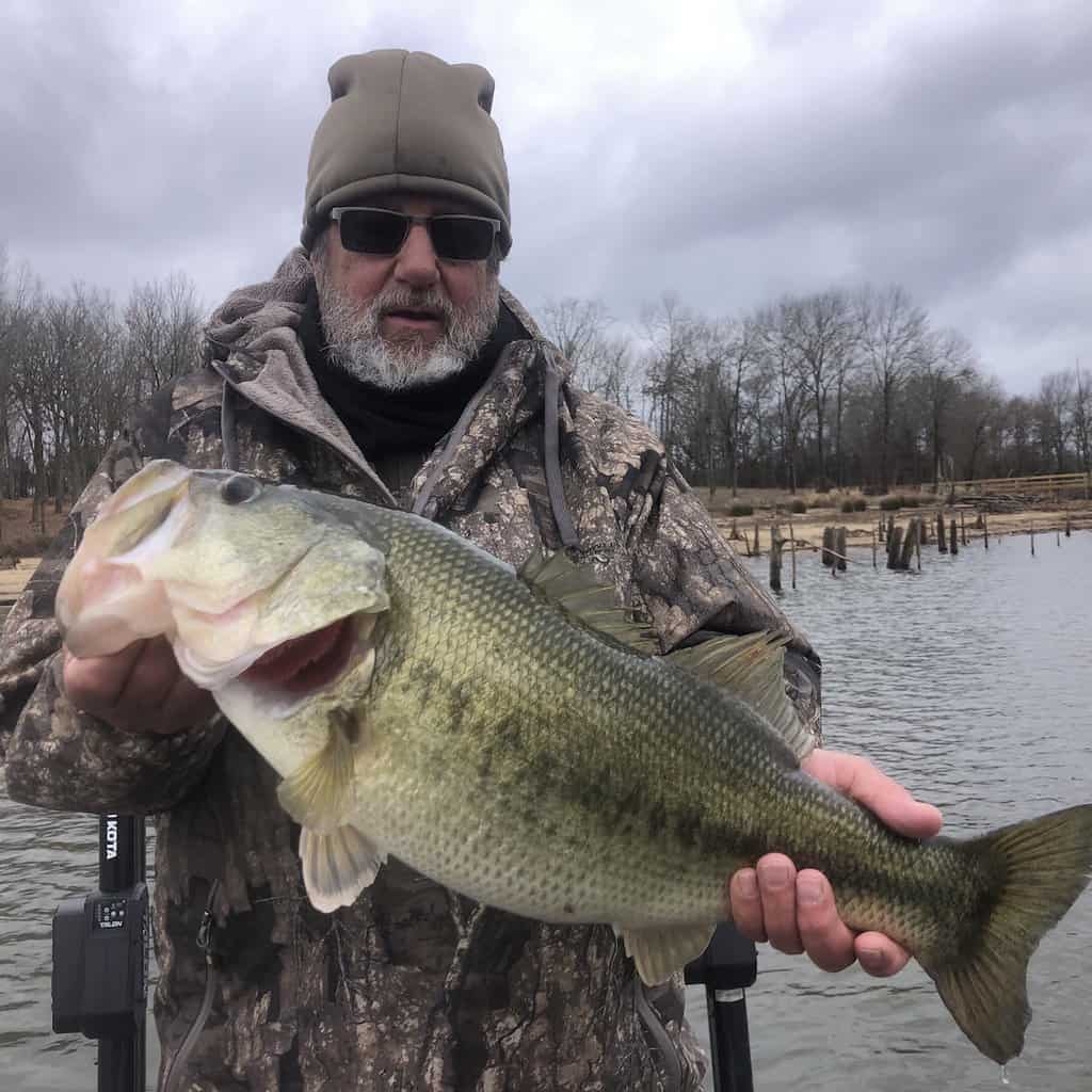 An angler dressed for cold weather holds up a fat largemouth bass with Lake Fork in the background.