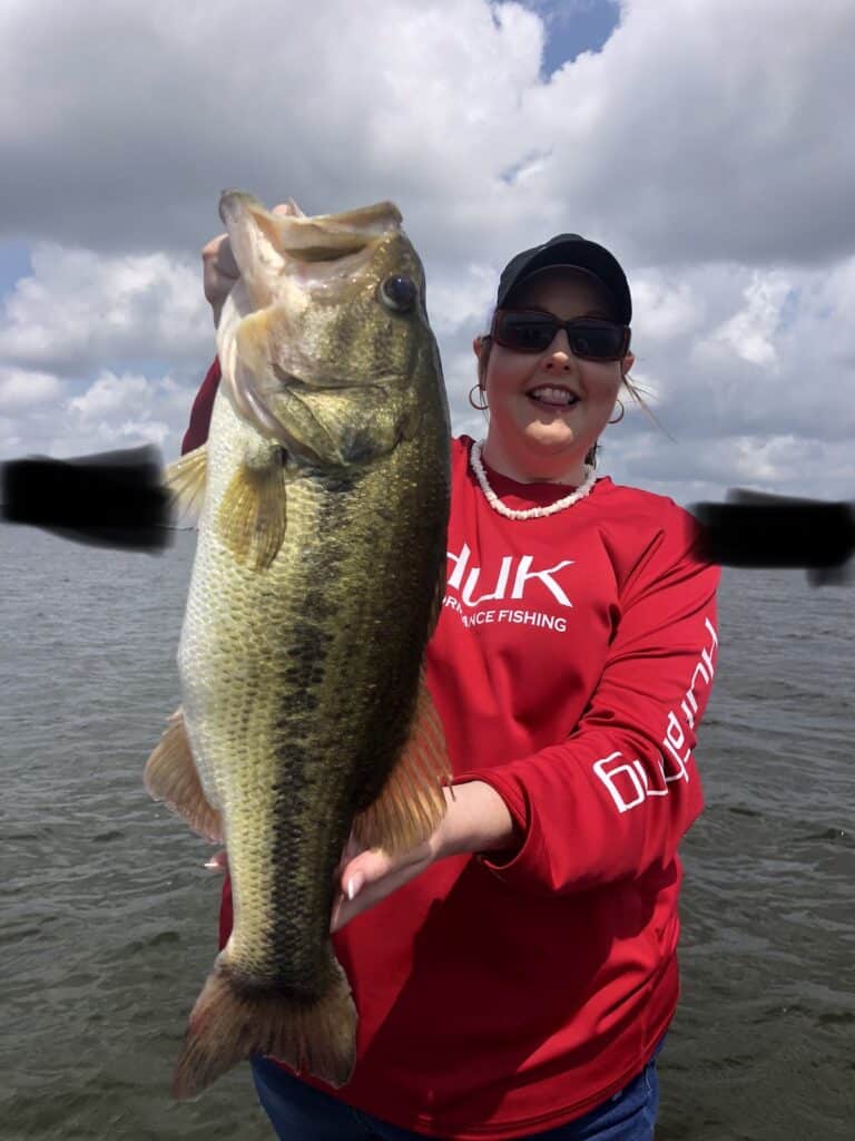 A woman in bright red holds up a large bass for a photo with the water of Lake Fork, Texas, behind the boat.
