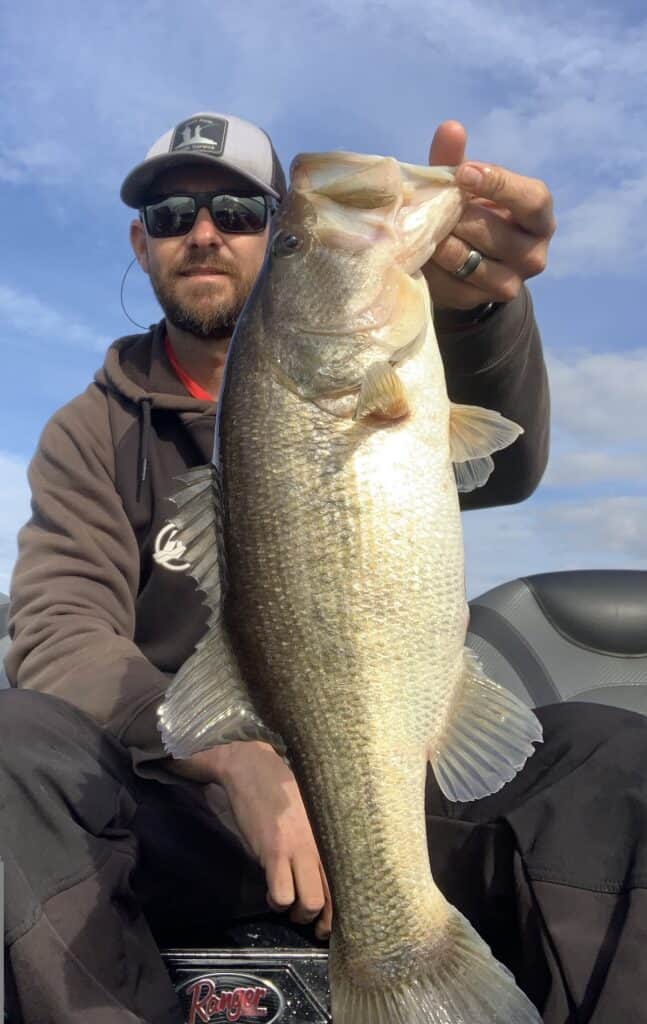 An angler holds up a large bass he caught fishing at Sam Rayburn Reservoir in Texas, with an outboard boat motor and mostly blue sky behind him.