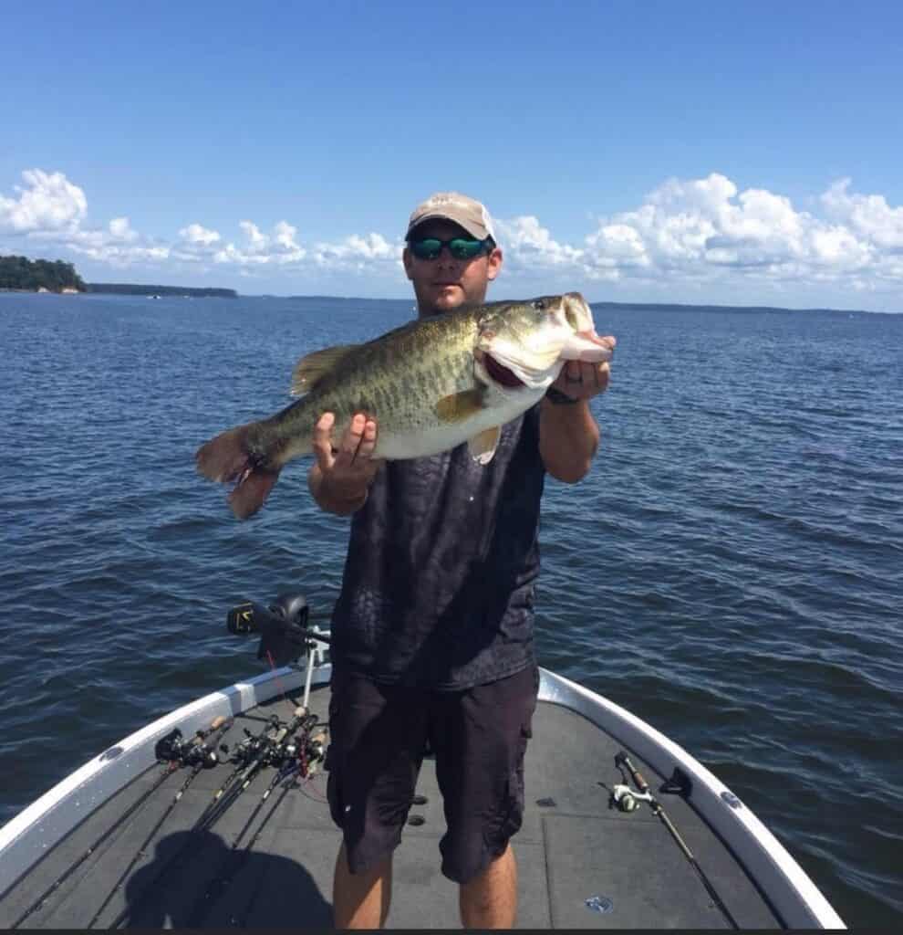 An angler standing at the front of a bass fishing boat holds a large bass he caught at Toledo Bend Reservoir, with the water behind him.