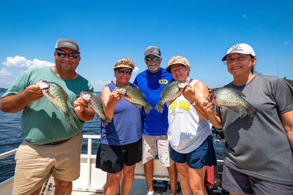 A group of four anglers and a guide standing on a boat hold up a bunch of crappie the caught fishing in Toledo Bend Reservoir, with the lake showing behind them.