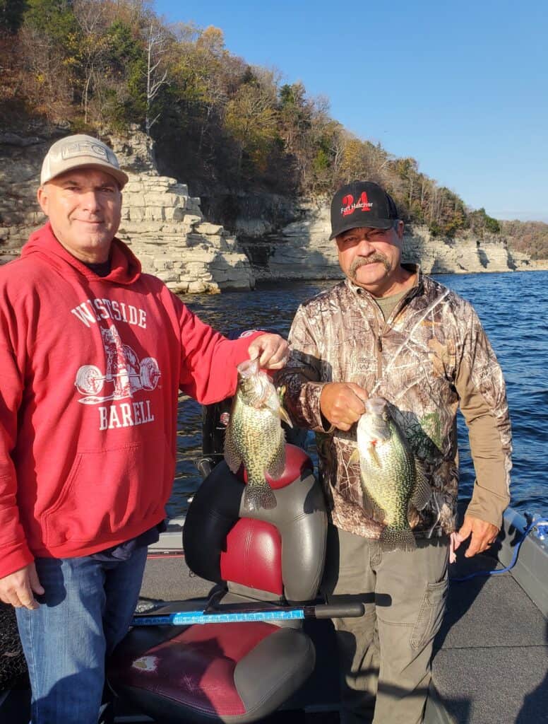 Two men hold up a crappie each standing on a boat with white rocky cliffs and dark blue water of Bull Shoals Lake in the background.