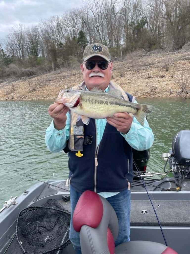 White mustached man in cap and sunglasses holds a largemouth bass he caught fishing in Bull Shoals Lake, with green water and barren shoreline in the background. 