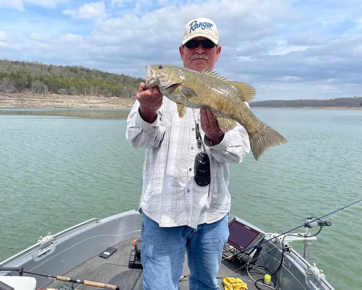 Angler in white cap standing in boat holds a large smallmouth bass caught fishing in Bull Shoals Lake, with the lake behind.