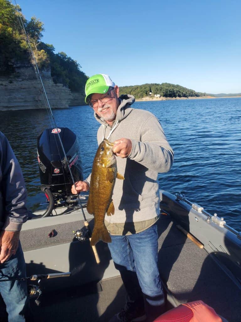 Smiling angler on a boat holds up a very olive colored smallmouth bass in one hand and fishing rod in the other, with cliffs and Bull Shoals Lake behind him.