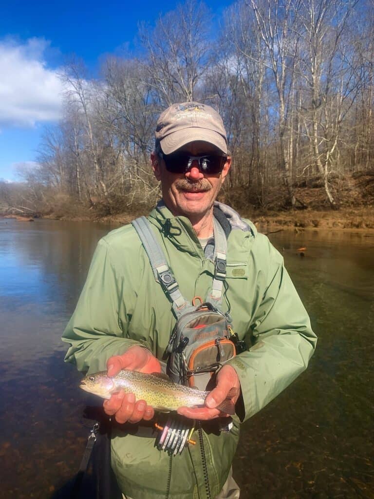 A fisherman wearing a ball cap, sunglasses and waders holds a rainbow trout with the waters of the Chattahoochee River and far bank behind him.