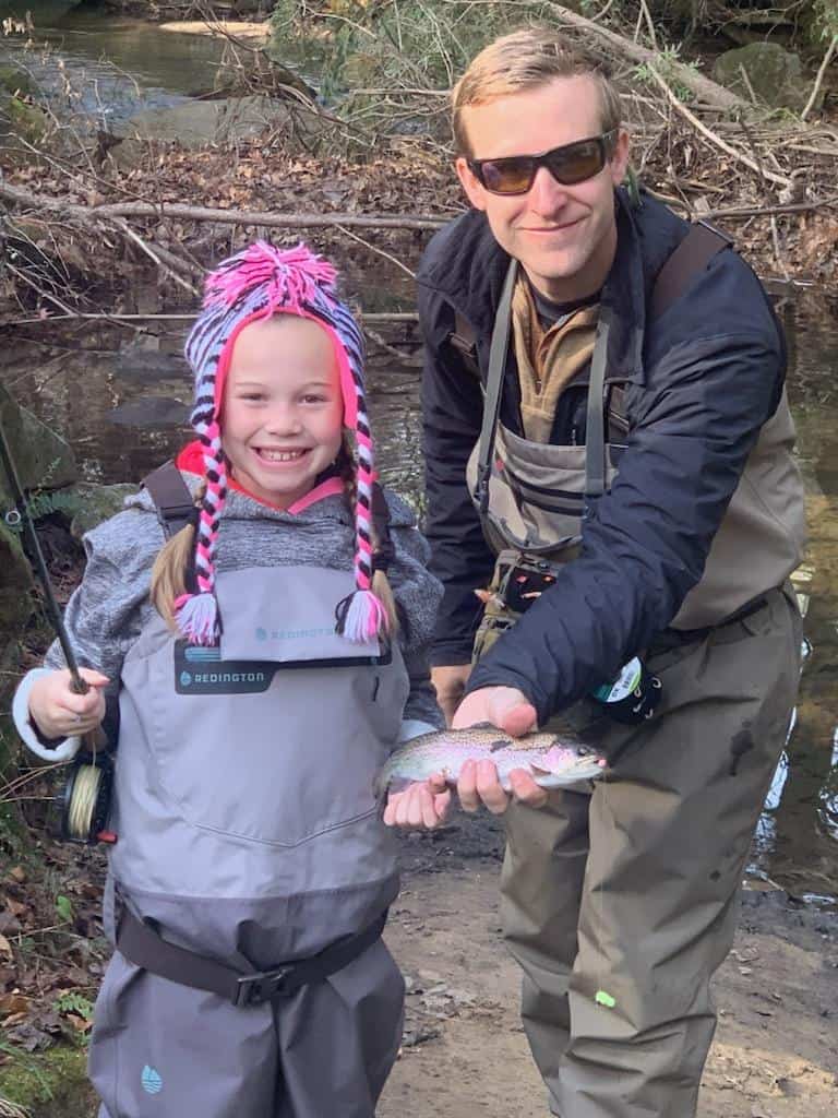 A young girl in a pink and purple knit cap smiles while a fishing guide holds a rainbow trout she caught fishing in Rock Creek, Georgia.