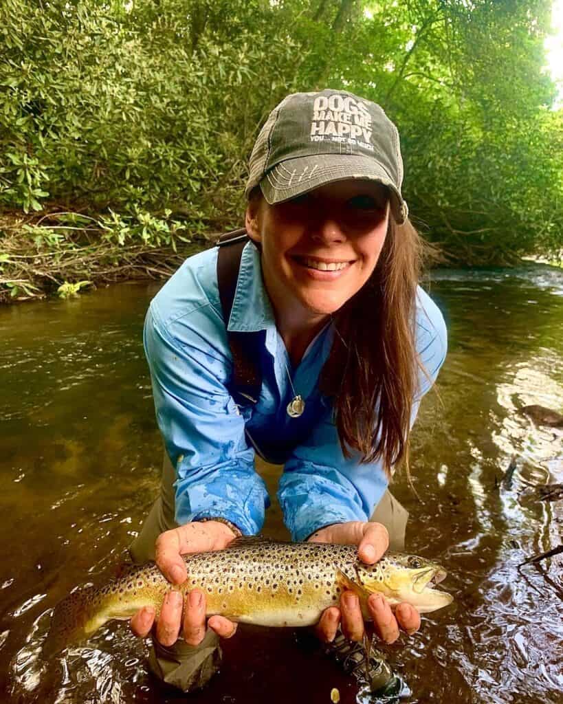 A woman wearing a ball cap that says 'Dogs Make Me Happy' and waders holds a brown trout while crouching down above Smith Creek.