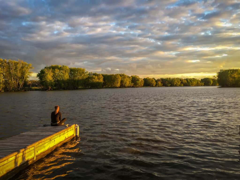 Woman sits on a dock looking out over the tree-lined shores of the Fox River, which is a fairly good place to catch walleye in Illinois.