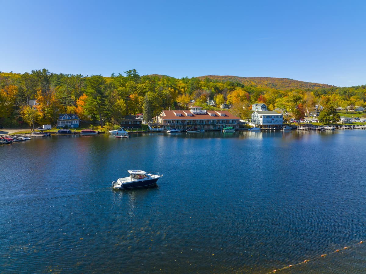 A white boat motors on the blue water of Lake Winnipesaukee in New Hampshire, with trees in background showing fall colors of yellow and orange.
