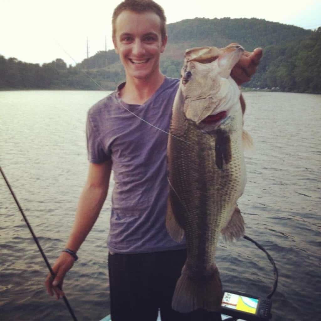 A young Alex Wetherell holds a massive largemouth bass in one hand and his fishing rod in the other while standing on a boat with Candelwood Lake in the background.