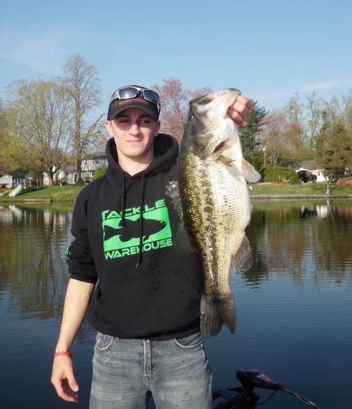 Alex Wetherell holds a trophy sized largemouth bass he caught fishing at Congamond Lakes in Connecticut, while standing on a boat with nice homes on the shore in the background.