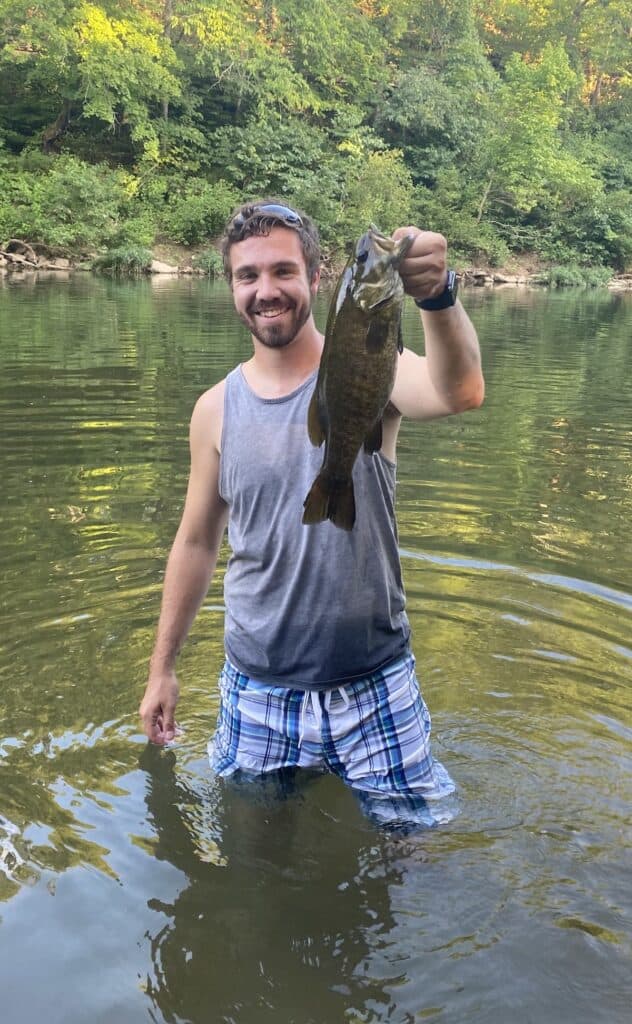 A smiling man wading in Missouri's Finley Creek holds a smallmouth bass.