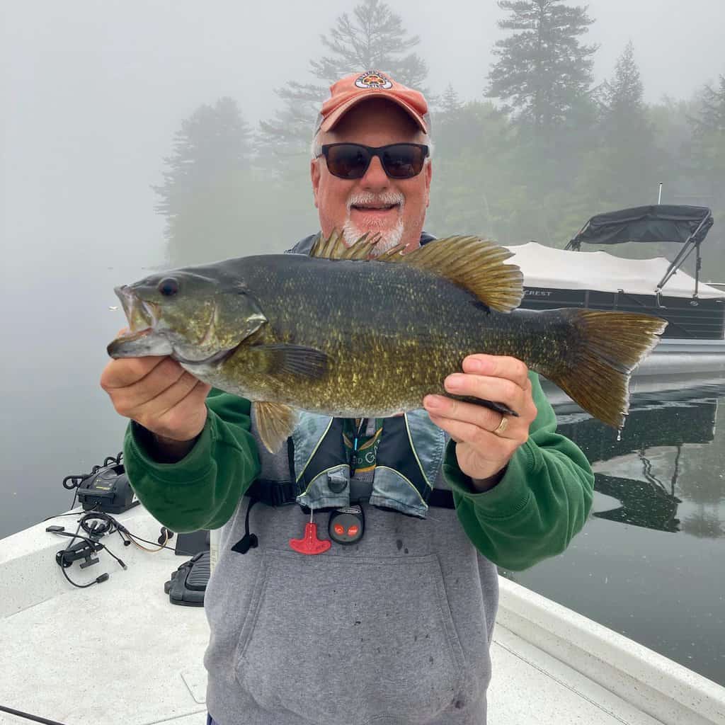 Smiling angler on a boat holds giant smallmouth bass caught fishing in the fog-shrouded lake behind him.