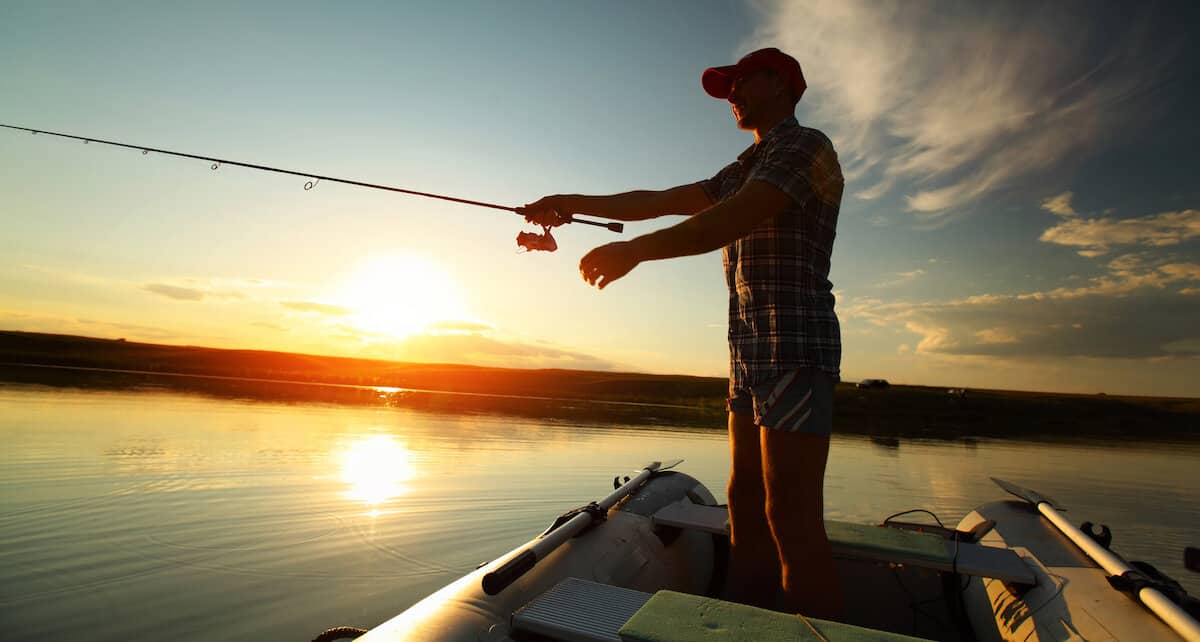 Man mostly in silhouette casting from a boat on a still lake at sunset.