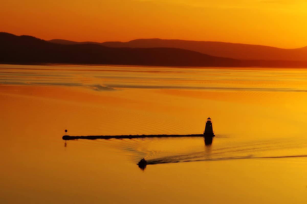 A fishing boat makes a wake near a lighthouse in a bright orange light of sunset on Lake Champlain.