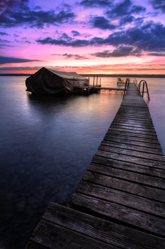 A beautiful autumn sunrise on the pebbled shores of Lake Cayuga in the Finger lakes region of New York state. A  wooden pier leads out to a power boat shelter and a deck with chairs for watching the sunrise.