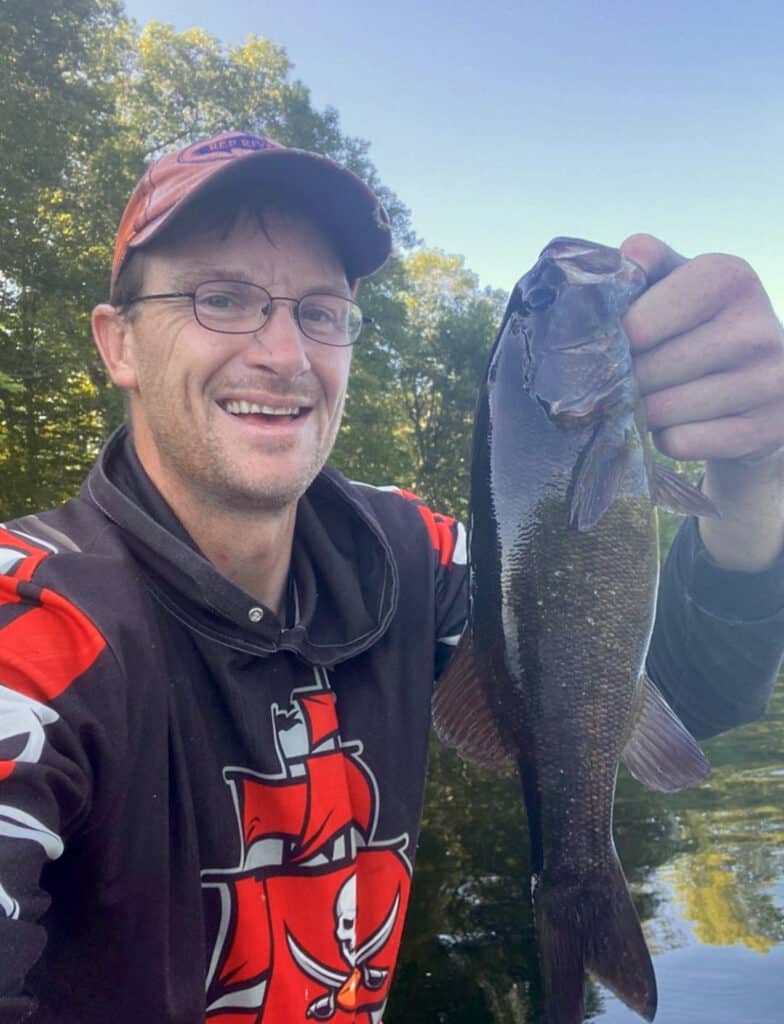 A smiling angler shows off a selfie while holding the lip of a smallmouth bass he caught fishing in Maine.