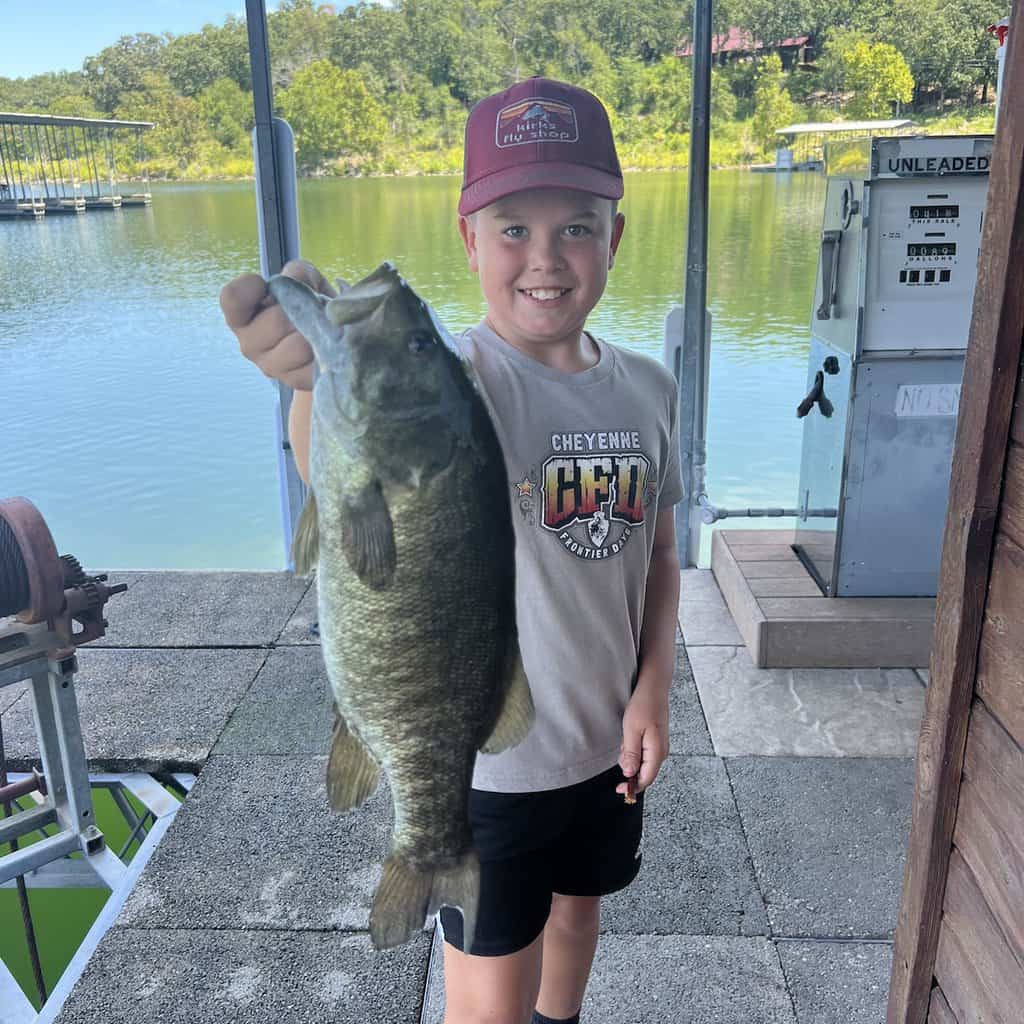 A boy on a dock holds up a very nice smallmouth bass he caught fishing at Tenkiller Lake in Oklahoma, with the lake in the background.