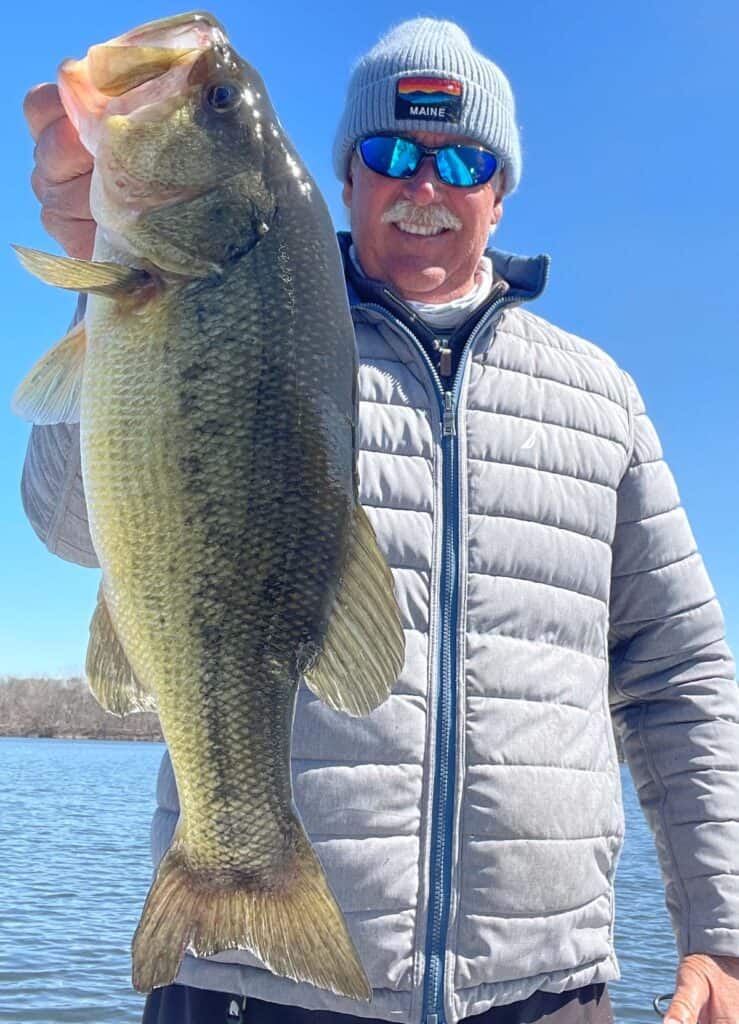 A smiling angler in reflective sunglasses holds a huge largemouth bass caught in Lake Champlain.