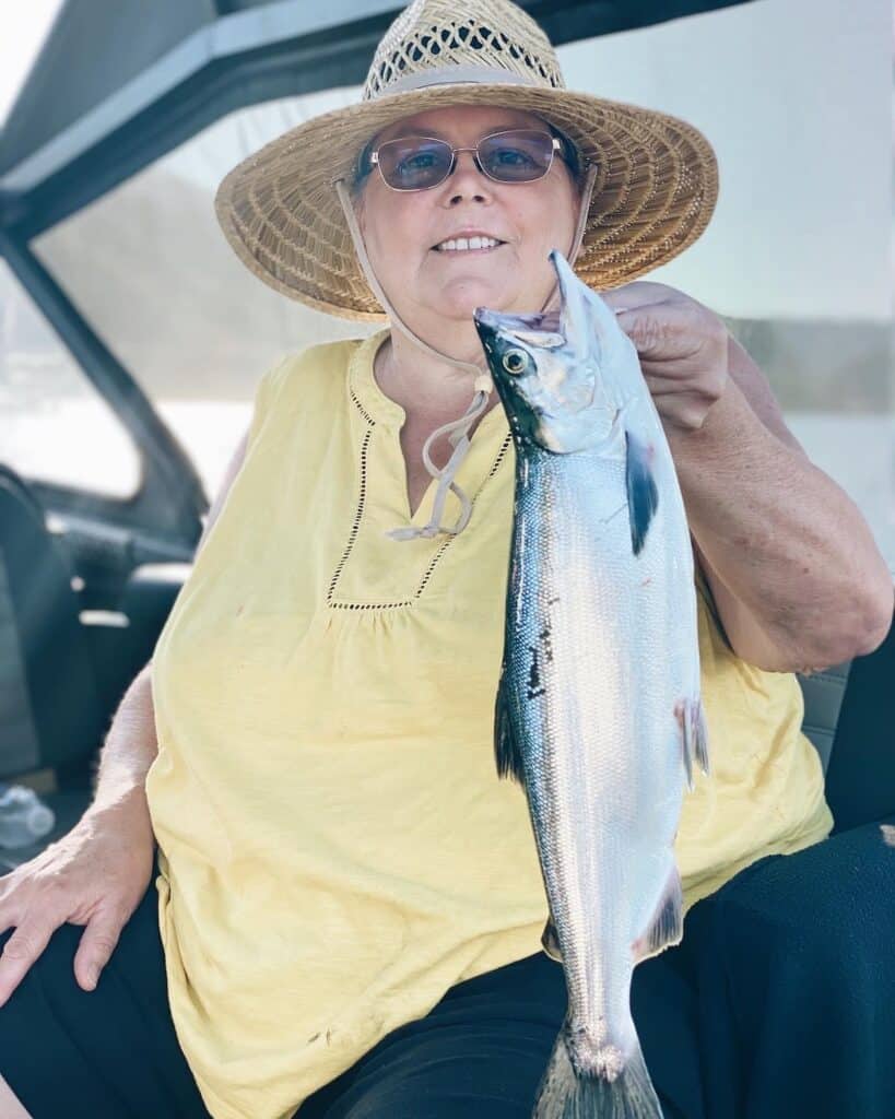 A smiling woman in a sun hat holds a large kokanee salmon she caught while fishing on a guide's boat at Whiskeytown Lake.