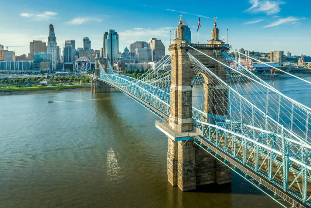 Panoramic view of downtown Cincinnati with a small fishing boat passing under the historic Roebling suspension bridge over the Ohio River.
