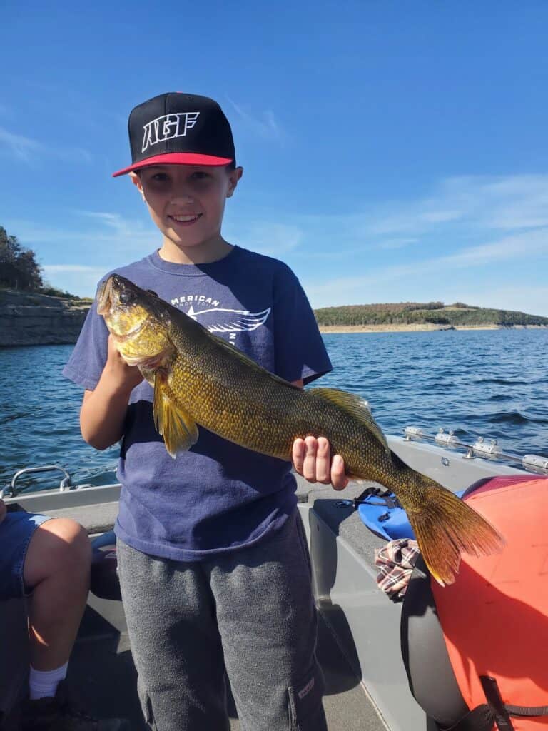 A young boy wearing a hat while on a boat smiles while holding a walleye he caught fishing at Bull Shoals Lake.