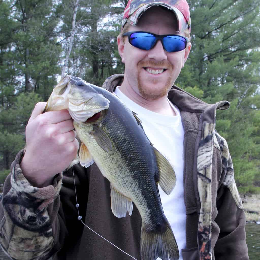 A smiling man wearing sunglasses holds up a largemouth bass he caught fishing.