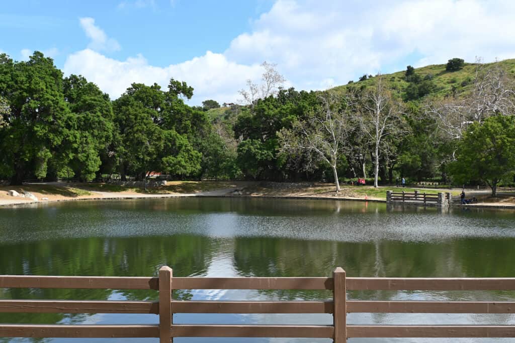 A fishing dock fence in the foreground and calm lake in the background at Irvine Regional Park's fishing pond.
