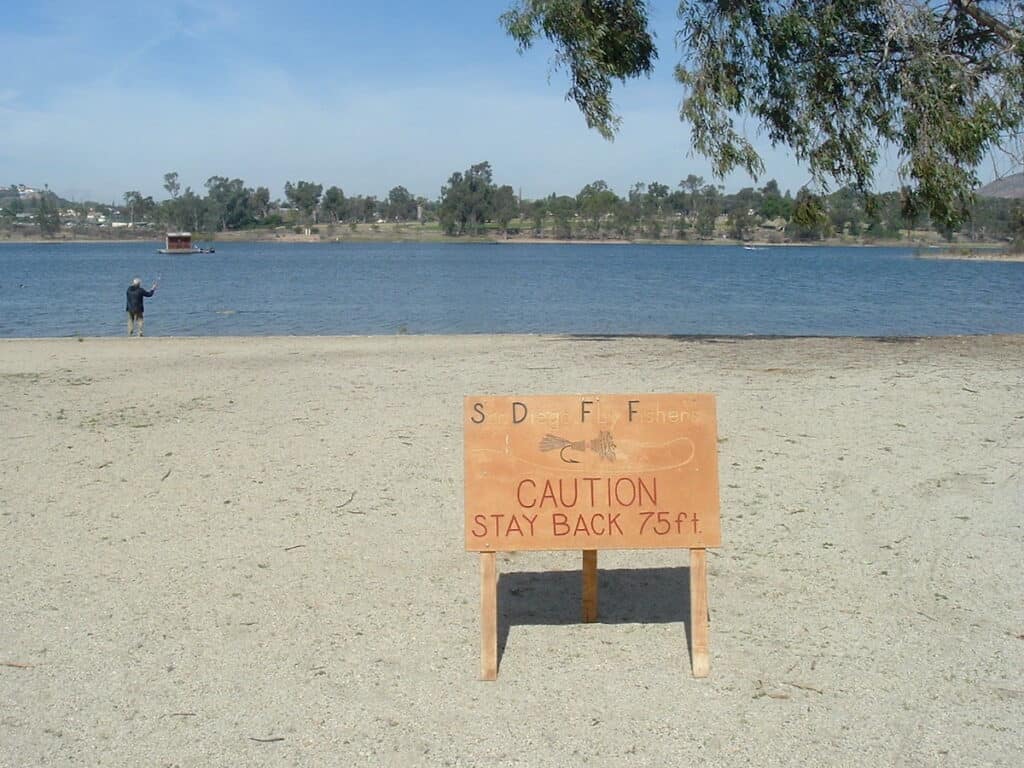 A caution sign warns passersby to stay back 75 feet as a fly angler casts from the sandy shoreline of Lake Murray.