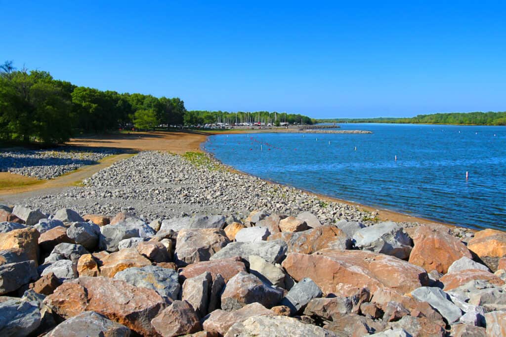 Rocky shoreline of Carlyle Lake can point to good fishing, with a swimming beach in the distance along the blue lake water.
