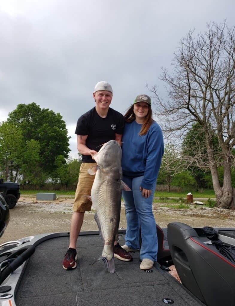 A young man and woman standing on a boat smile as they hold up a giant catfish they caught fishing at Truman Lake in Missouri.