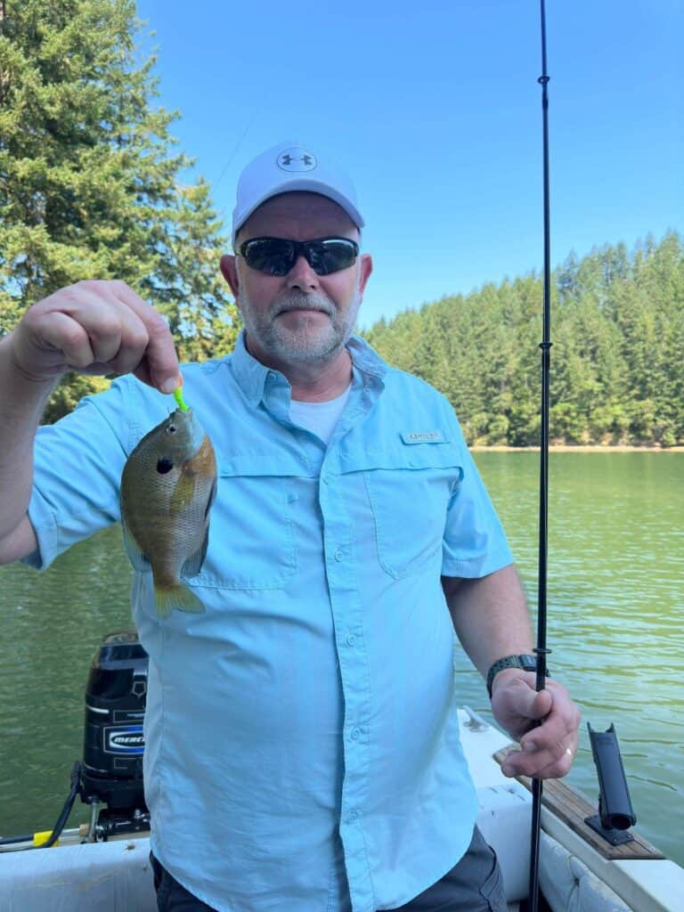 Angler holds a bluegill with a chartreuse jig in its mouth with Cooper Creek Reservoir in the background.