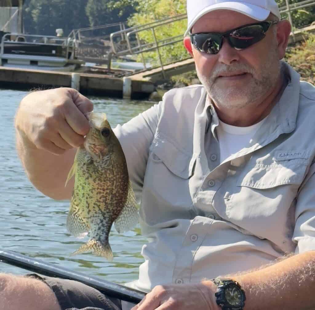 A man in sunglasses sitting in a boat holds up a fair-sized crappie with docks and water of Loon Lake, Oregon, in the background.