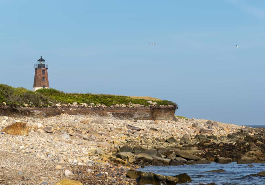 Heavily eroded beach of sand and stone beneath the Point Judith Lighthouse in Narragansett, Rhode Island, with a blue sky background.