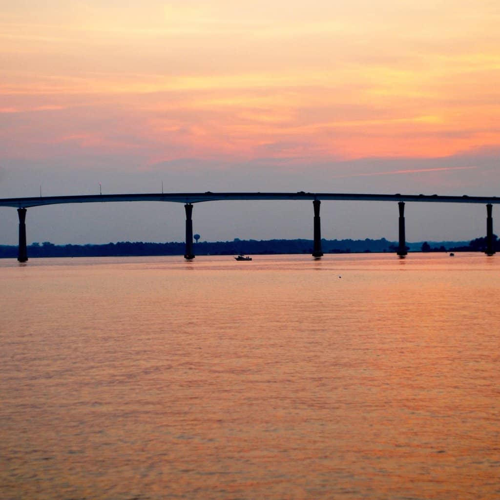 Orange sunset with the Chesapeake Bay Bridge and Tunnel with fishing boats beneath it.