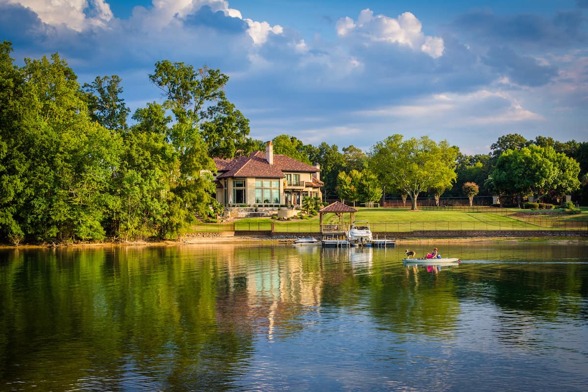 A man and a dog in a small boat passing a huge home and boat dock along the shore of Lake Norman.