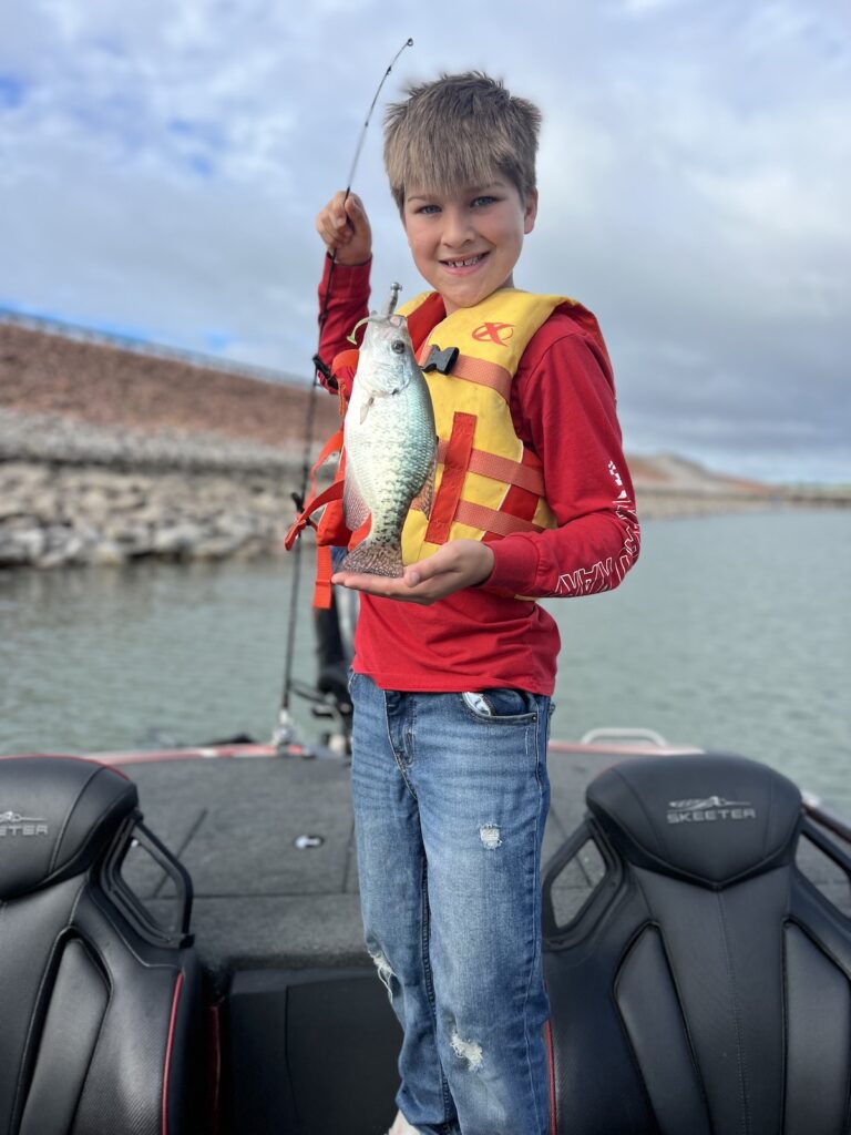 A smiling boy on a boat holds up a crappie still hooked on a jig with a rocky riprap bank in the background at Canton Lake, Oklahoma.