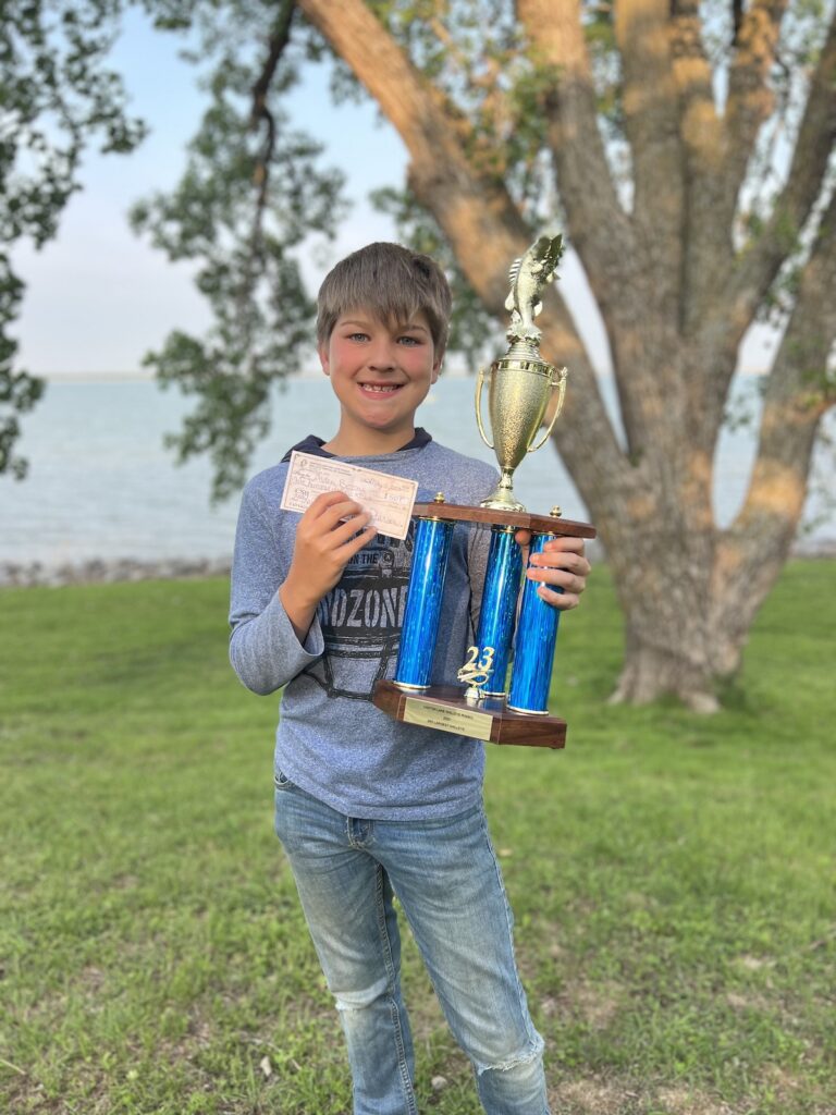 A boy holds a large trophy and a hefty check he won in the Canton Lake Walleye Rodeo, with Canton Lake in the background.
