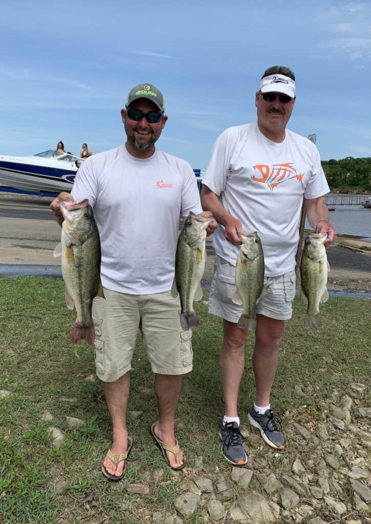 Two anglers each hold a pair of largemouth bass they caught fishing at Eufaula Lake, with a boat on a ramp at lakeside behind them.