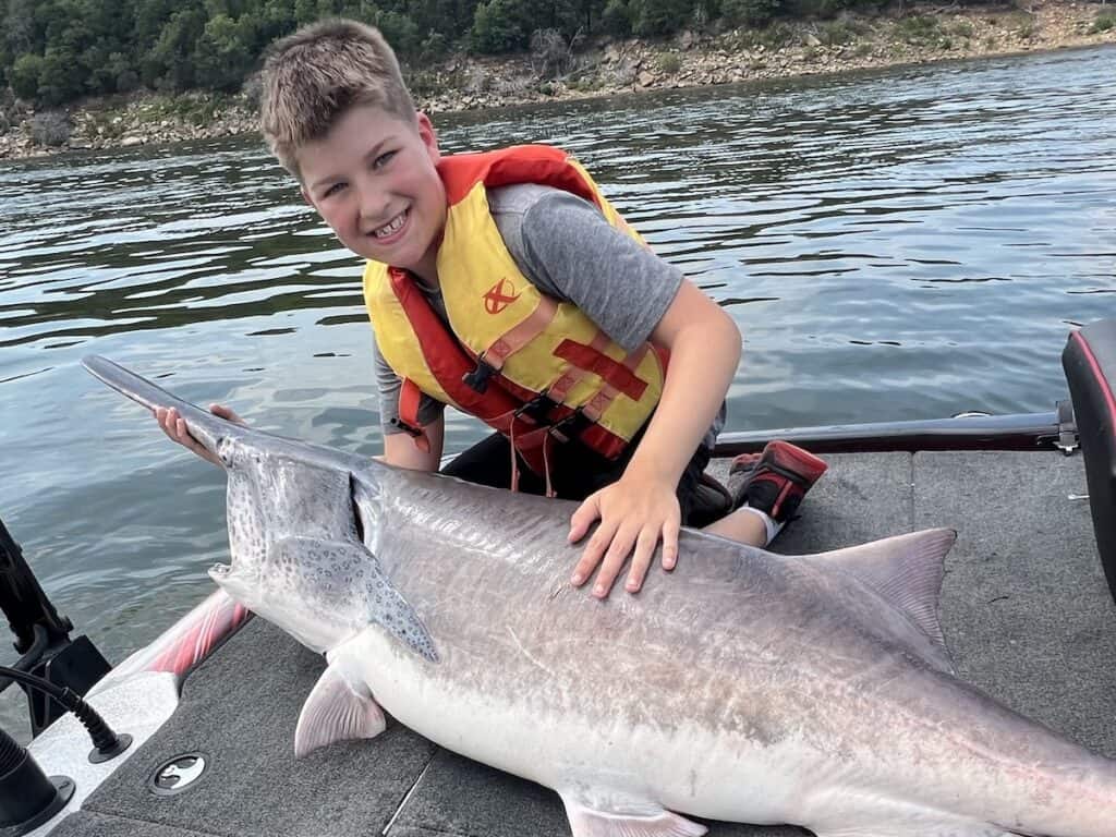 A smiling boy holds a spoonbill (paddlefish) by the bill on the deck of a fishing boat on Keystone Lake, Oklahoma.